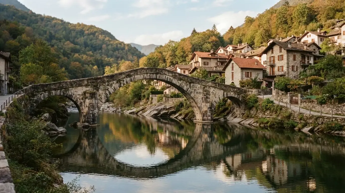 Eine alte Steinbrücke über einen ruhigen Fluss, dahinter Häuser mit Ziegeldächern und bewaldete Hügel.