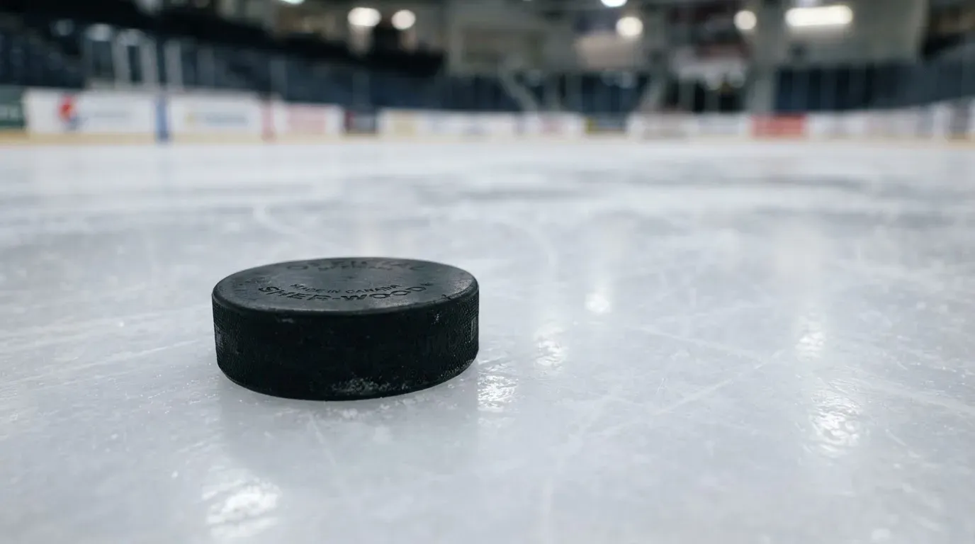 Ein Eishockeypuck liegt auf dem Eis, im Hintergrund unscharf die Bande unter hellem Hallenlicht.