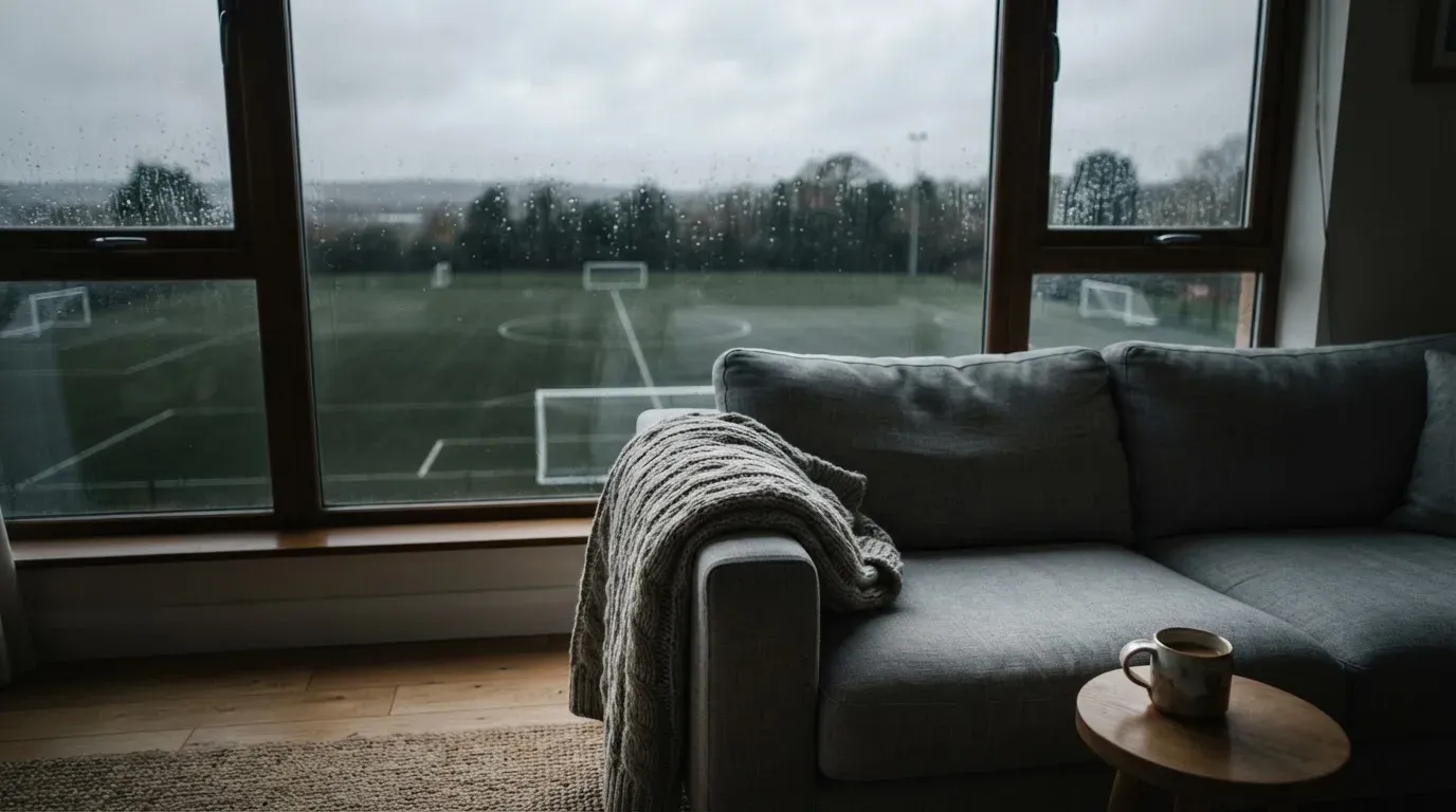 Ein Sofa mit Wolldecke steht vor einem grossen Fenster, draussen Regen und in der Ferne ein Fussballplatz.