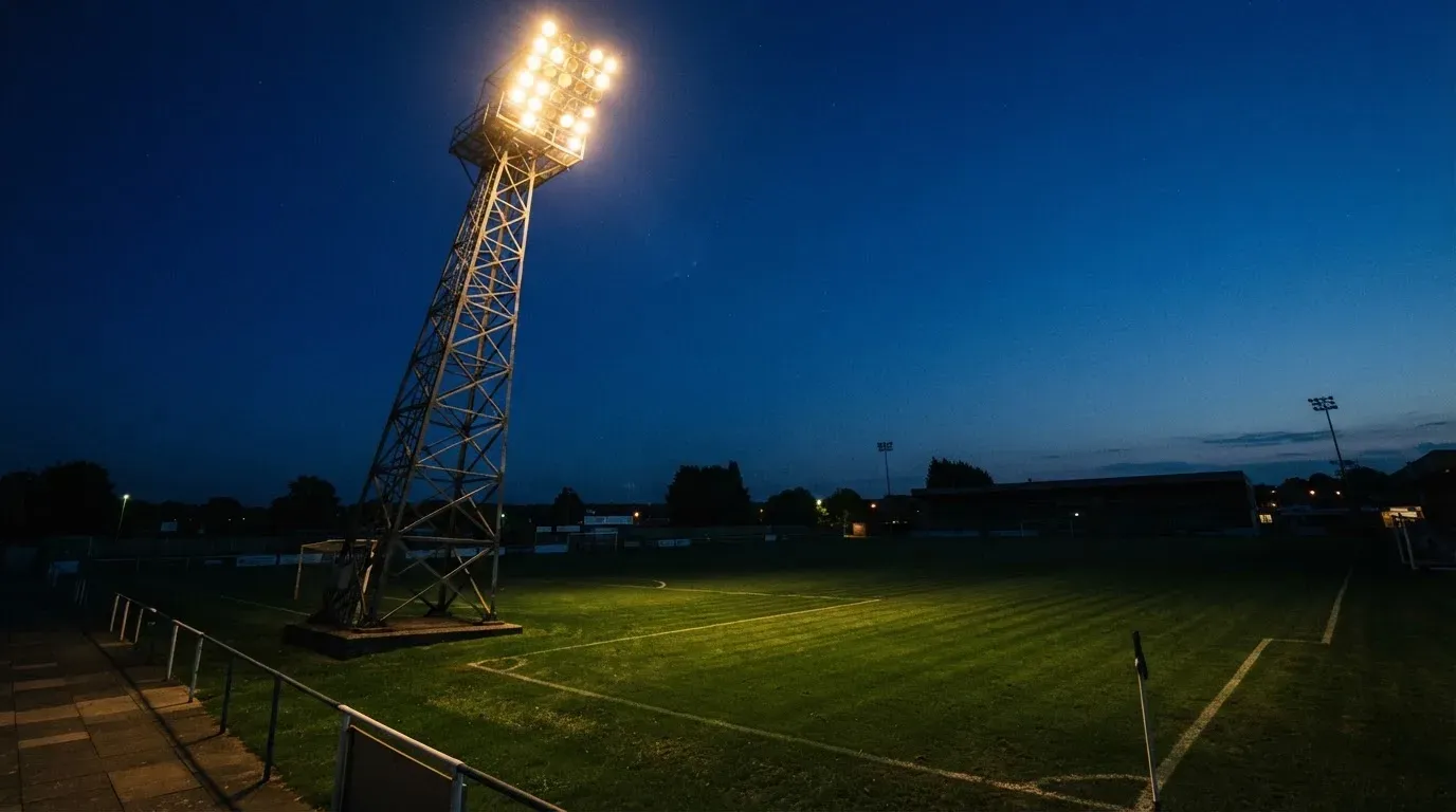 Ein Flutlichtmast ragt in den Abendhimmel, die Rasenfläche darunter liegt leer und dunkelgrün da.