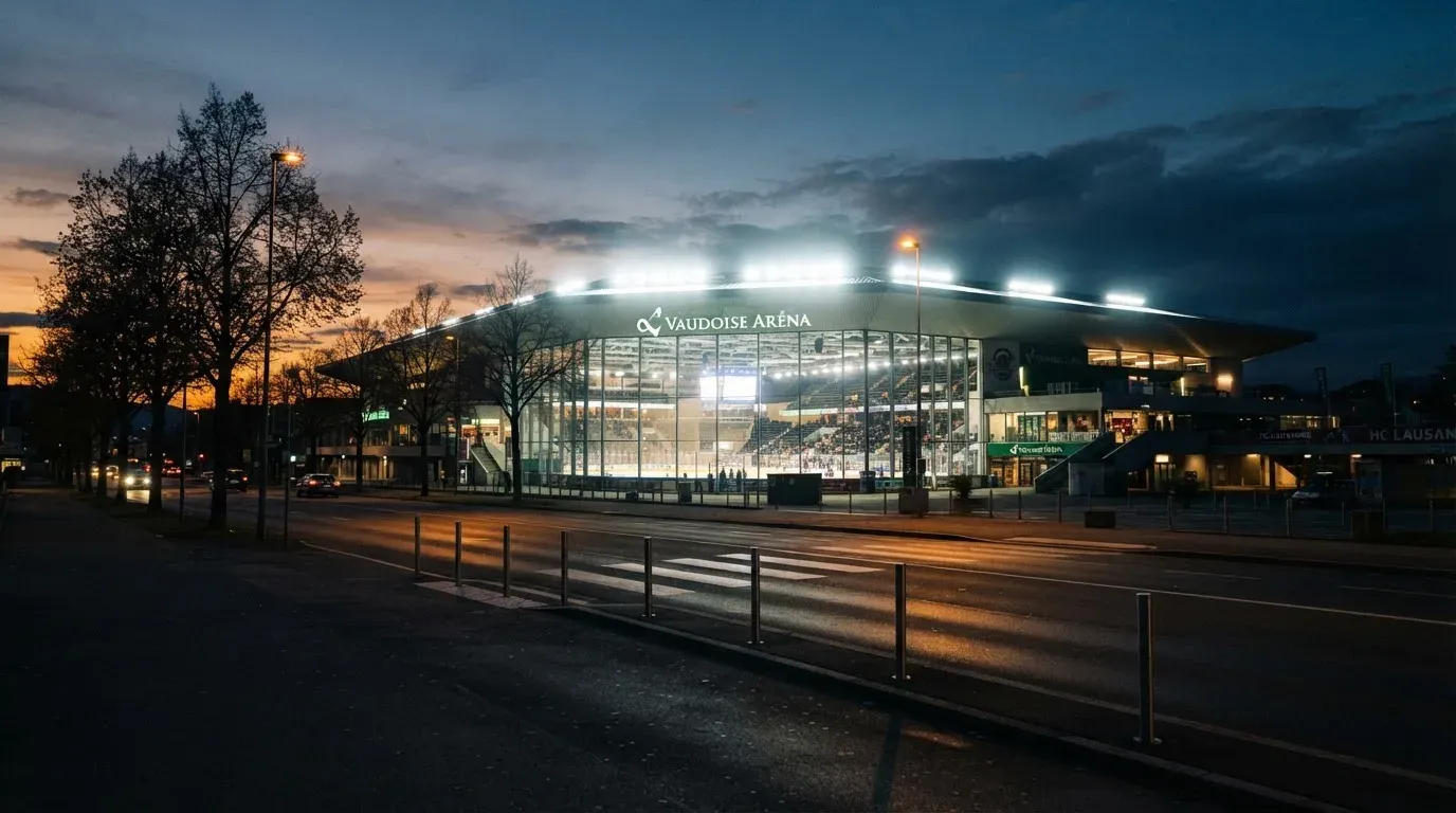 Ein grosses Eisstadion aus der Ferne gesehen, hell erleuchtet von Flutlicht in der Abenddämmerung, davor eine leere Strasse.