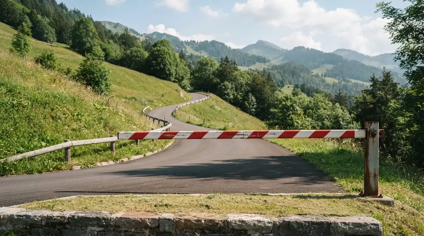 Ein Schlagbaum sperrt eine Landstrasse in den Bergen, dahinter windet sich der Weg zwischen grünen Hängen.