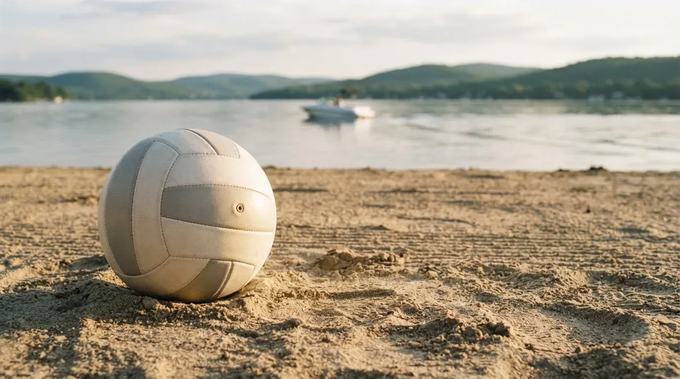 Ein Volleyball liegt im Sand eines Spielfelds am Seeufer, im Hintergrund gleitet ein Boot über das Wasser.