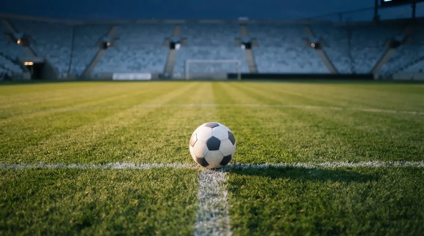 Ein Fussball liegt auf dem Mittelkreis eines leeren Rasenplatzes, dahinter ragen leere Tribünenreihen in den Abendhimmel.