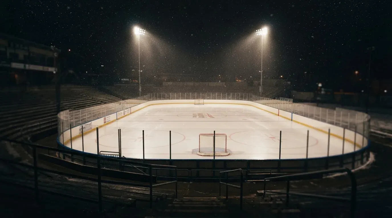 Eine Eishockeybahn liegt unter Flutlicht aus der Ferne betrachtet, leichter Schneefall treibt durch den Lichtkegel über dem Eis.