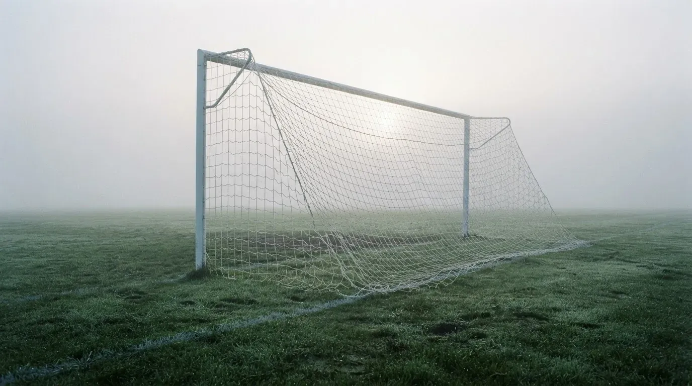 Eine Torlinie mit Netz steht am frühen Morgen im dichten Nebel auf einem Rasenplatz, die Umrisse der Pfosten verschwimmen.