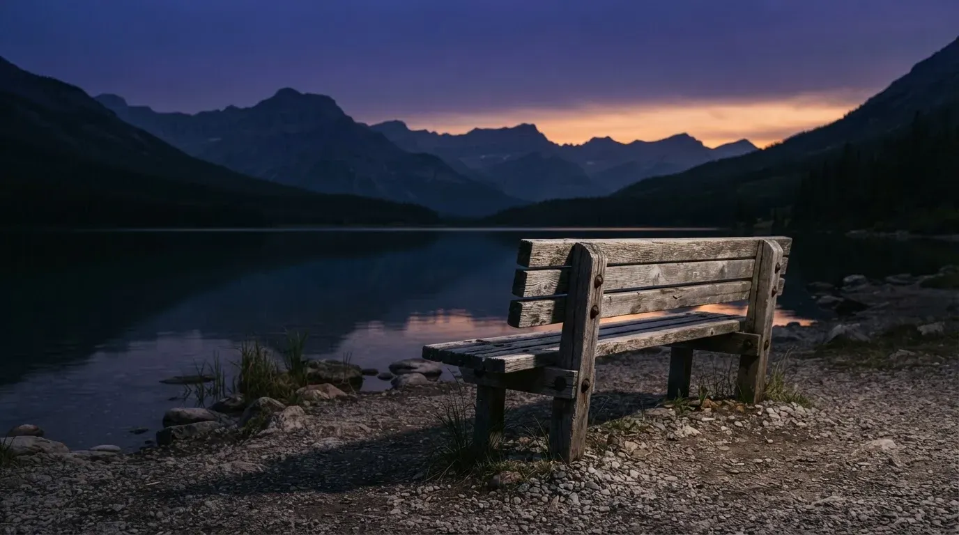 Eine Holzbank steht in der Dämmerung am Seeufer, im Hintergrund zeichnet sich ein Bergpanorama gegen den violetten Himmel ab.