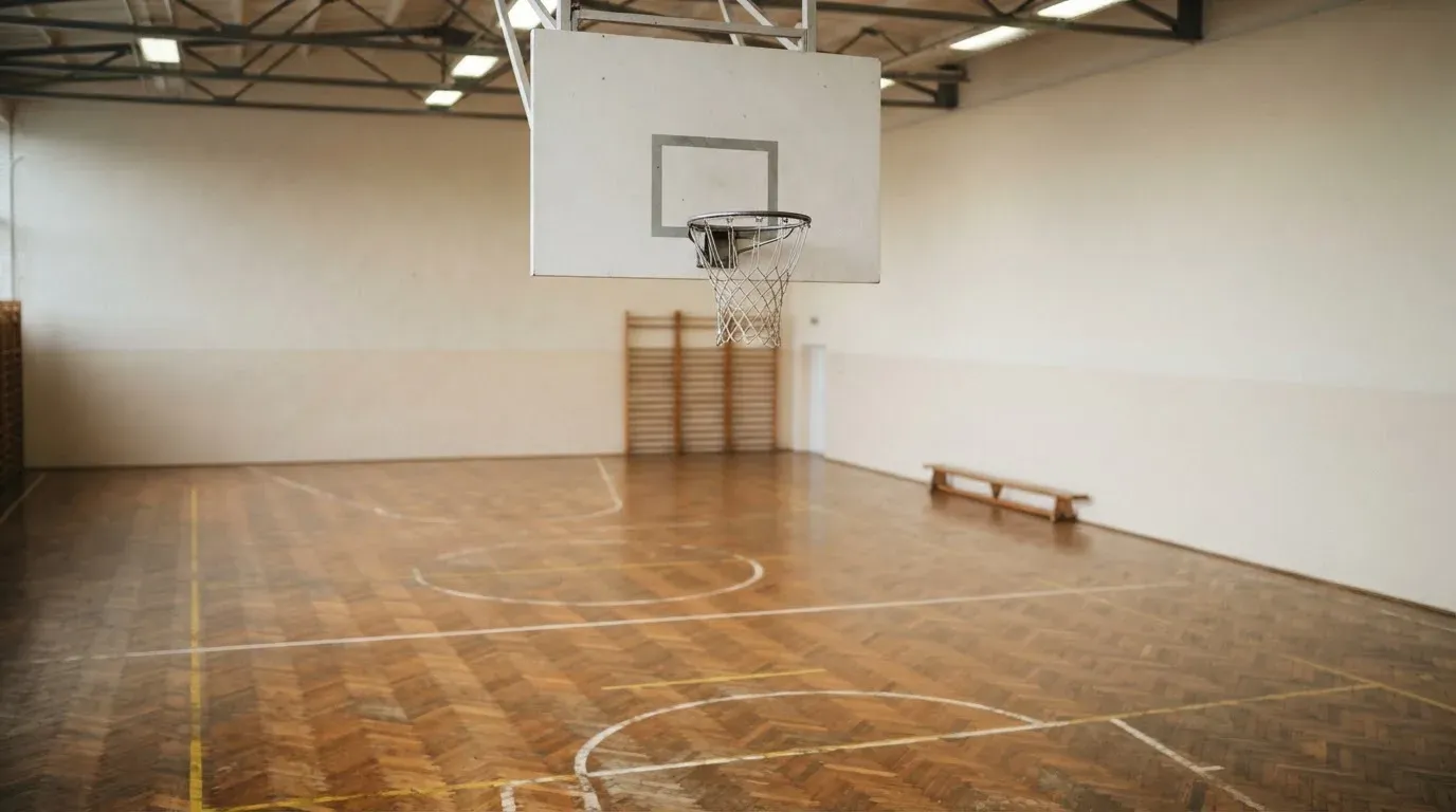 Ein Basketballkorb hängt in einer leeren Halle über einem Holzparkett, von der Decke fällt gleichmässiges Licht auf den Boden.