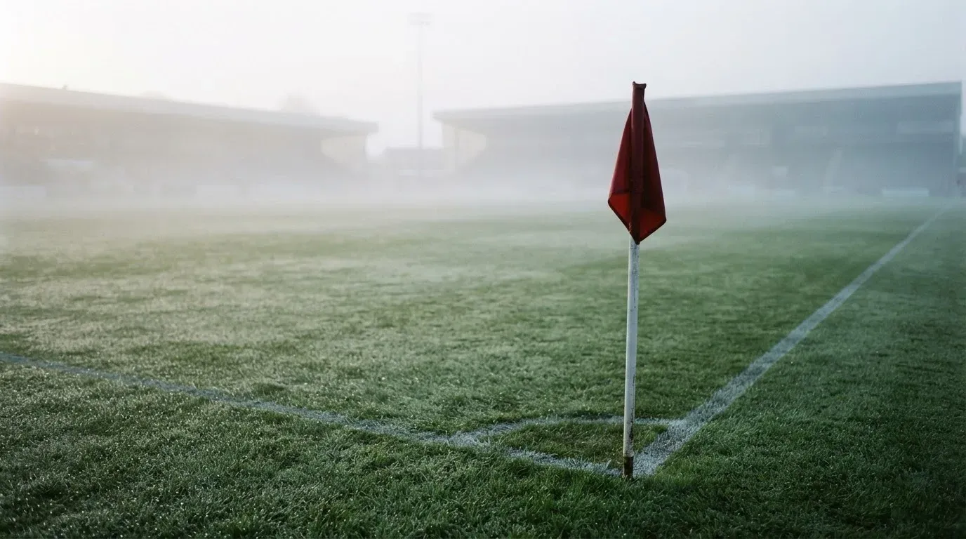 Eine Eckfahne steht am Morgen auf nassem Rasen, Tautropfen hängen im Gras und über dem Platz liegt leichter Dunst.