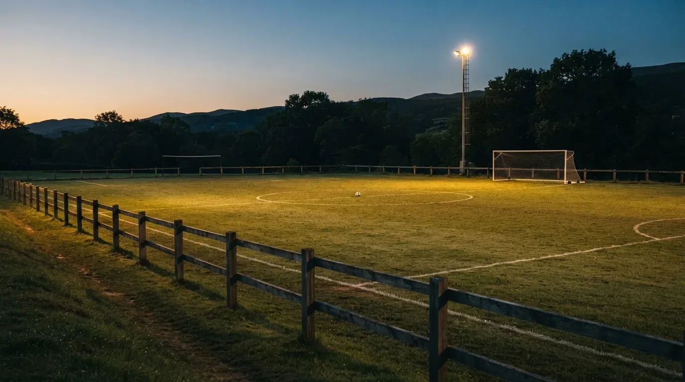 Ein kleiner Dorf-Fussballplatz liegt in der Dämmerung unter einem hohen Flutlichtmast, am Spielfeldrand steht ein Holzzaun.