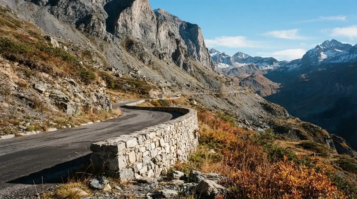 Eine Passstrasse im Hochgebirge säumt eine niedrige Steinmauer, dahinter ragen Felswände auf und der Herbsthimmel ist klar.