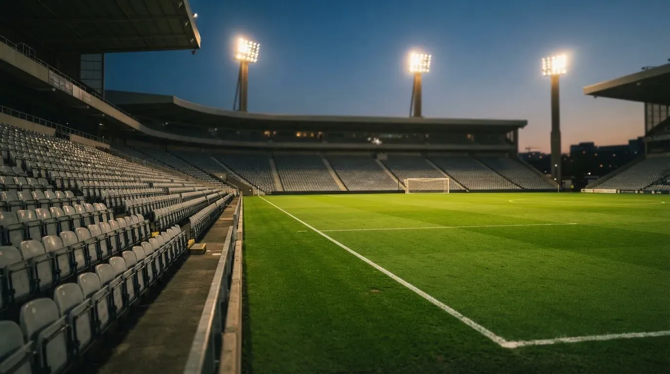Leere Tribünenränge eines Fussballstadions in der Abenddämmerung, über dem Rasen leuchten die Flutlichter.
