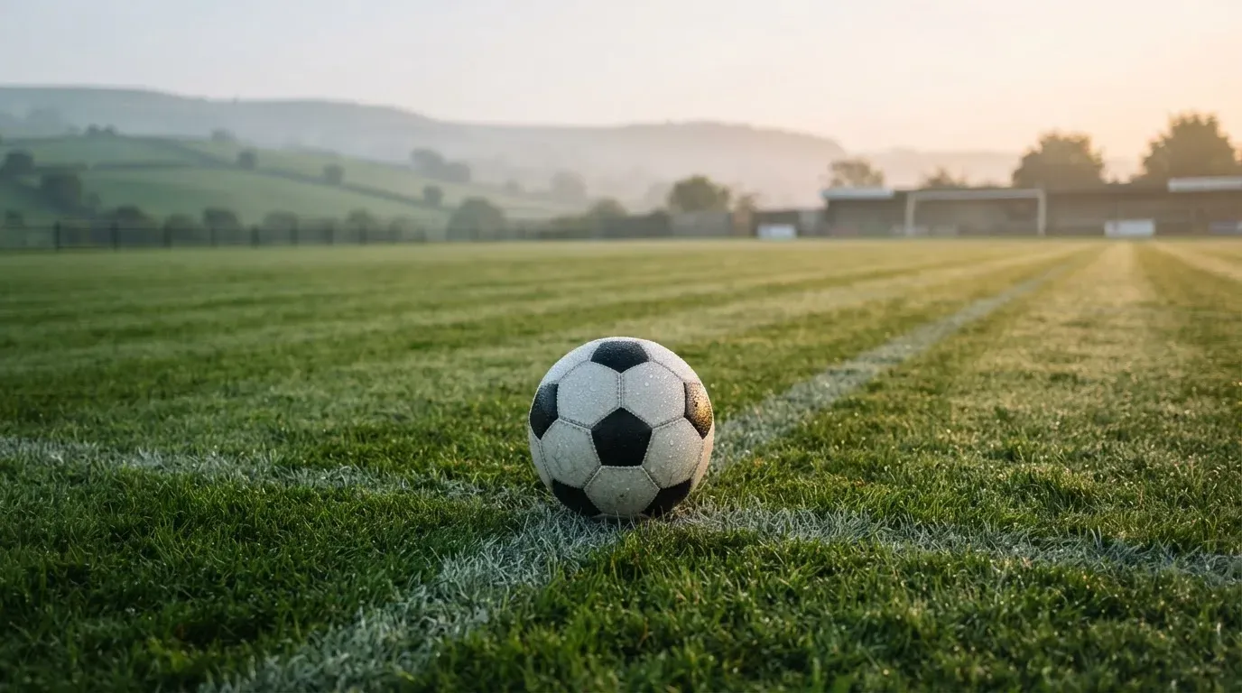 Ein Fussball liegt am frühen Morgen auf dem Anstosspunkt eines taunassen Rasens, im Hintergrund erheben sich Hügel im Dunst.