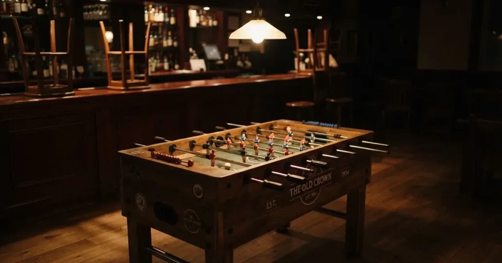 Ein hölzerner Tischfussball steht in einer leeren Bar, die Stangen mit den Figuren glänzen im Gegenlicht einer Lampe.