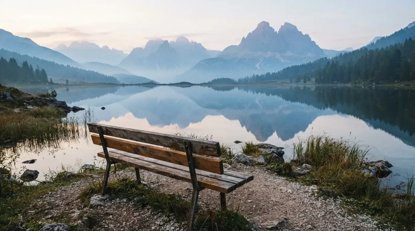 Eine Holzbank an einem Seeufer mit Blick auf eine Bergkette im Morgendunst, das Wasser ist spiegelglatt.