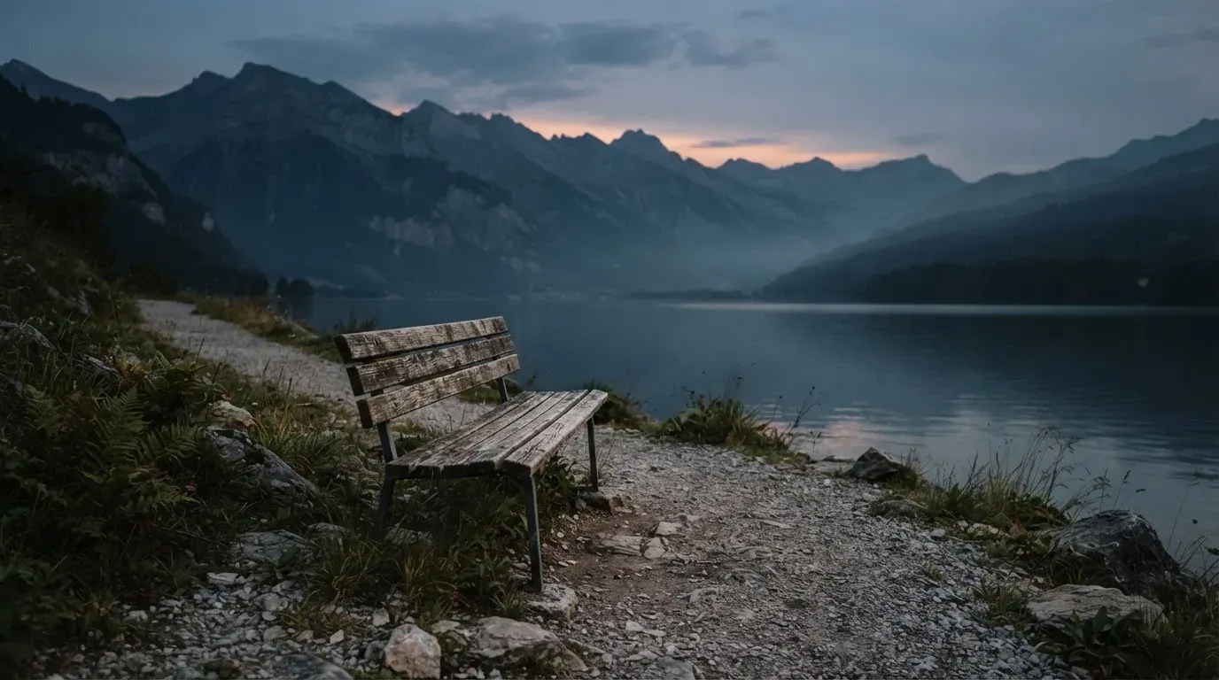 Eine leere Holzbank am Seeufer in der Dämmerung, im Hintergrund dunkle Berge