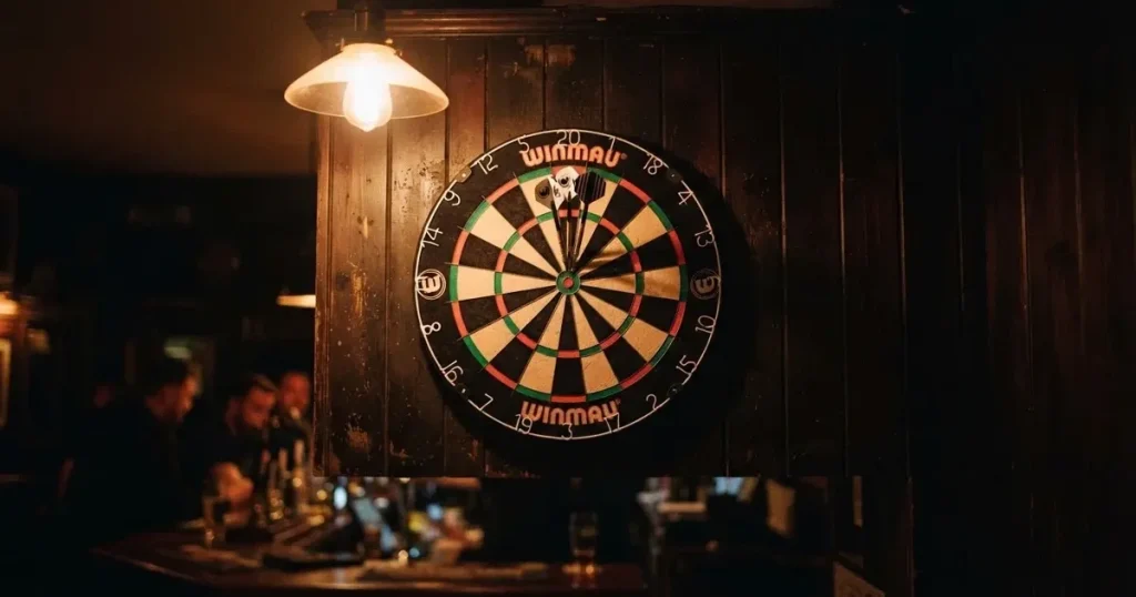 Drei Dartpfeile stecken in einer Dartscheibe an einer Holzwand in einer dämmrigen Kneipe, warmes Lampenlicht fällt von oben