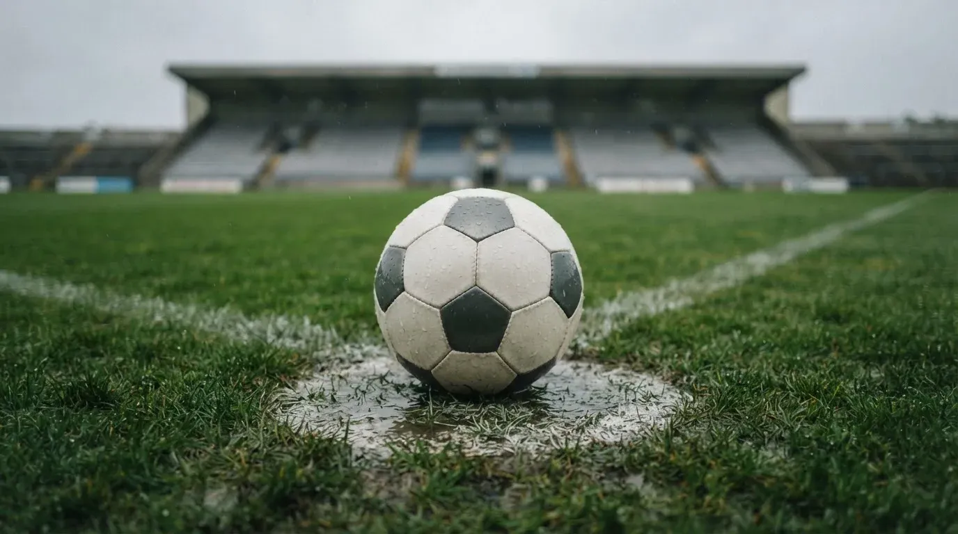 Ein Fussball liegt auf dem Anstosspunkt eines nassen Rasens, dahinter eine leere Tribüne unter grauem Himmel.