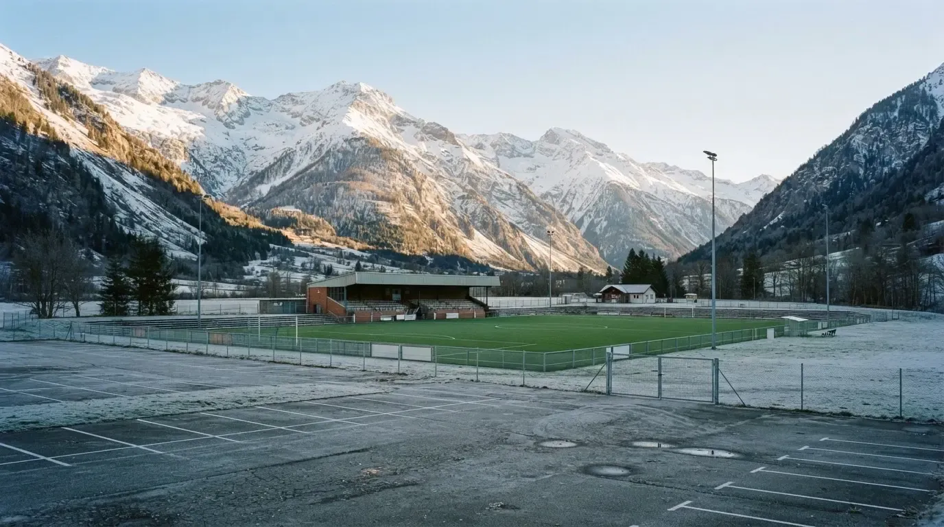 Schneebedeckte Berge ragen hinter einem kleinen Stadion in einem Alpental auf, im Vordergrund ein leerer Parkplatz am Morgen.