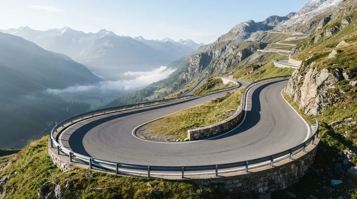 Eine Passstrasse in den Alpen mit enger Kurve und Leitplanke, darüber ein klarer Morgenhimmel und einzelne Nebelschwaden im Tal.