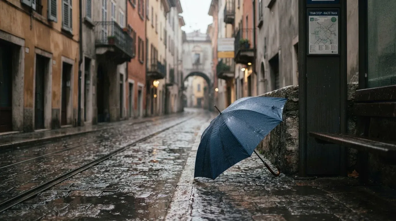 Eine Tramhaltestelle bei Regen in einer Altstadtgasse, das Kopfsteinpflaster glänzt und ein einzelner Regenschirm lehnt an einem Mäuerchen.