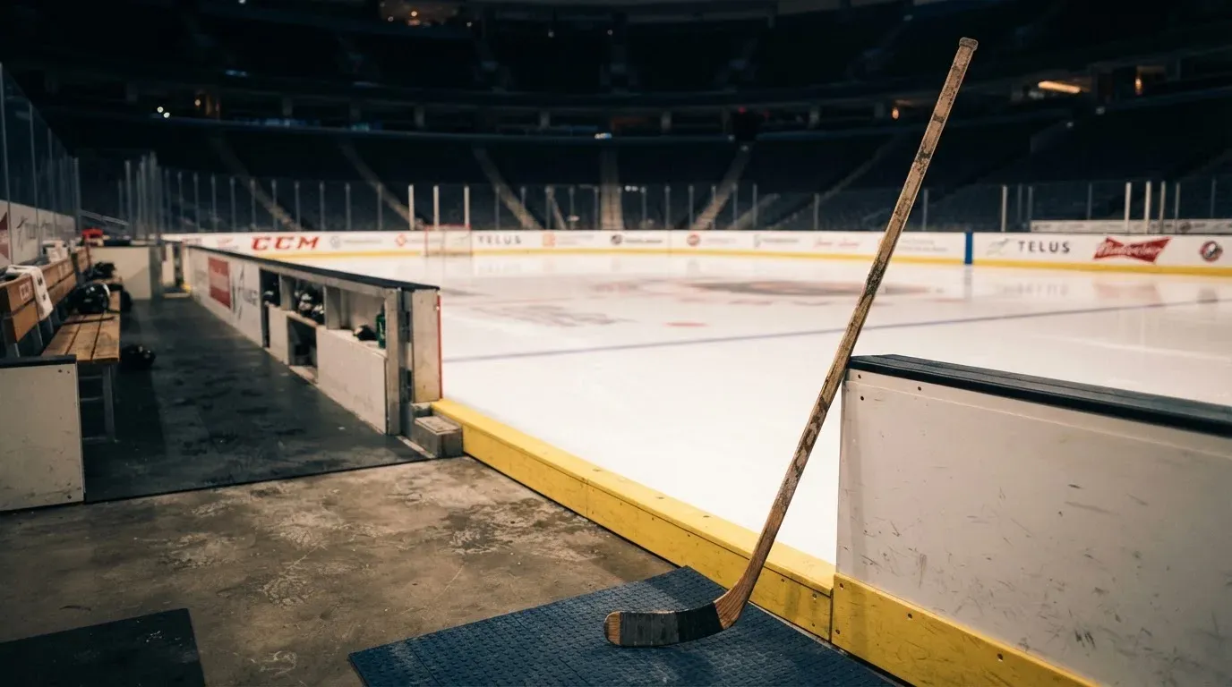 Ein Eishockeyschläger lehnt an der Bande, in der Ferne liegt die hell beleuchtete Eisfläche einer Halle unter Flutlicht.