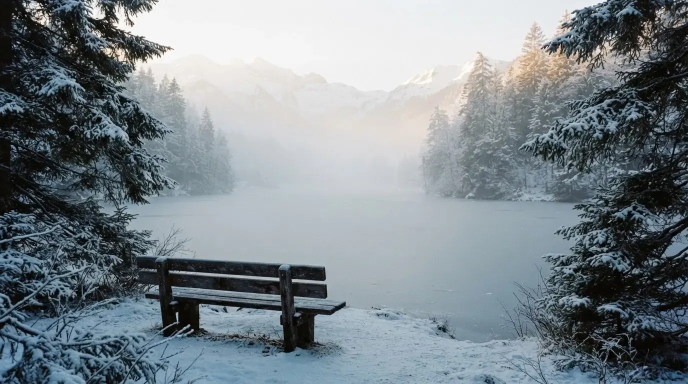 Ein zugefrorener Bergsee im Morgendunst, am Ufer steht eine einsame Holzbank zwischen verschneiten Tannen.