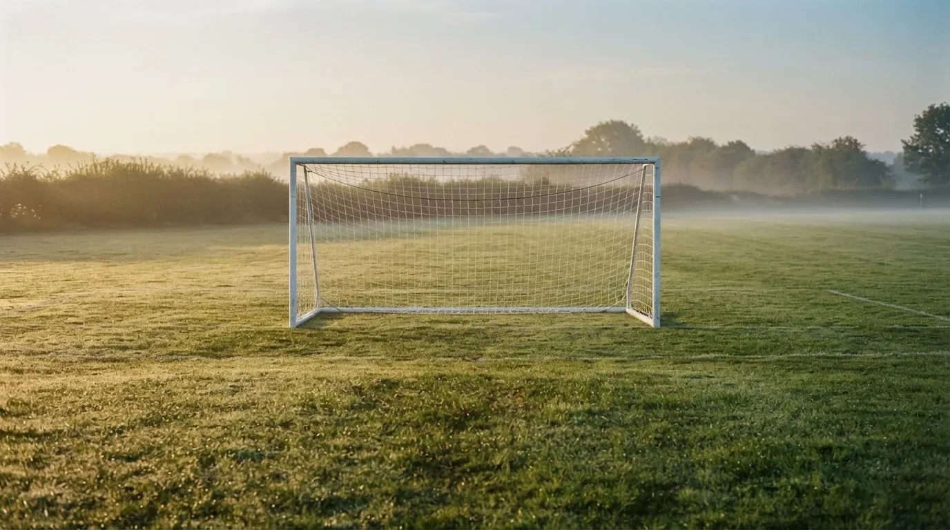 Ein Fussballtor mit Netz in der Totalen auf einem leeren Platz, Tau glänzt im Gras und die Morgensonne steht tief.