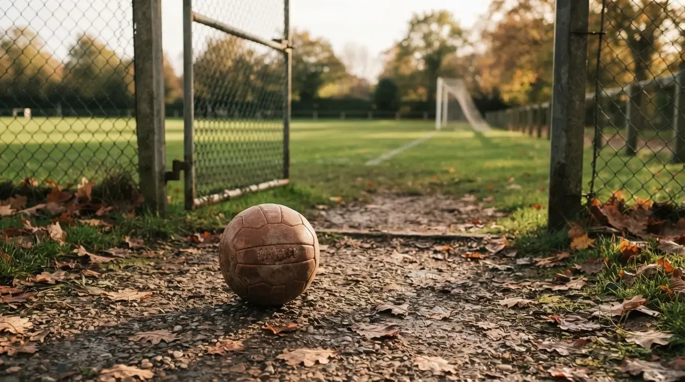 Ein abgenutzter Fussball liegt auf einem Kiesweg neben dem Eingang eines Vereinsplatzes, Laub liegt auf dem Boden.