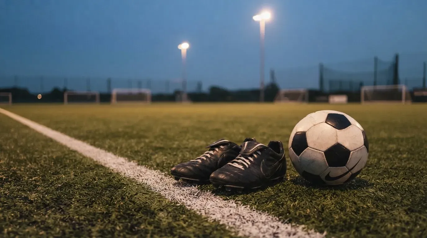Ein Fussball und ein Paar Stollenschuhe stehen am Rand eines Kunstrasenplatzes, Flutlichtmasten ragen in den Abendhimmel.