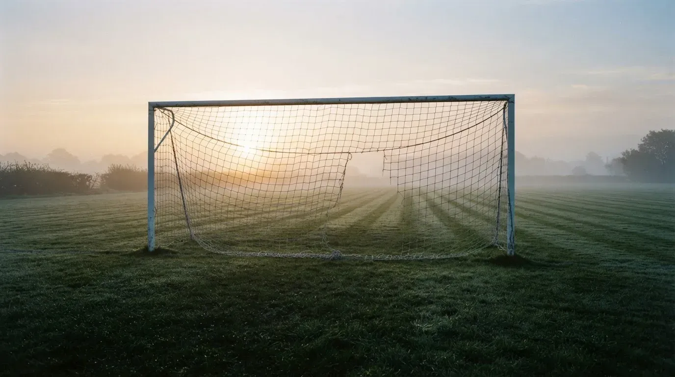 Ein Torpfosten mit Netz steht im Gegenlicht auf einem leeren Fussballplatz, über dem Rasen liegt Nebel.