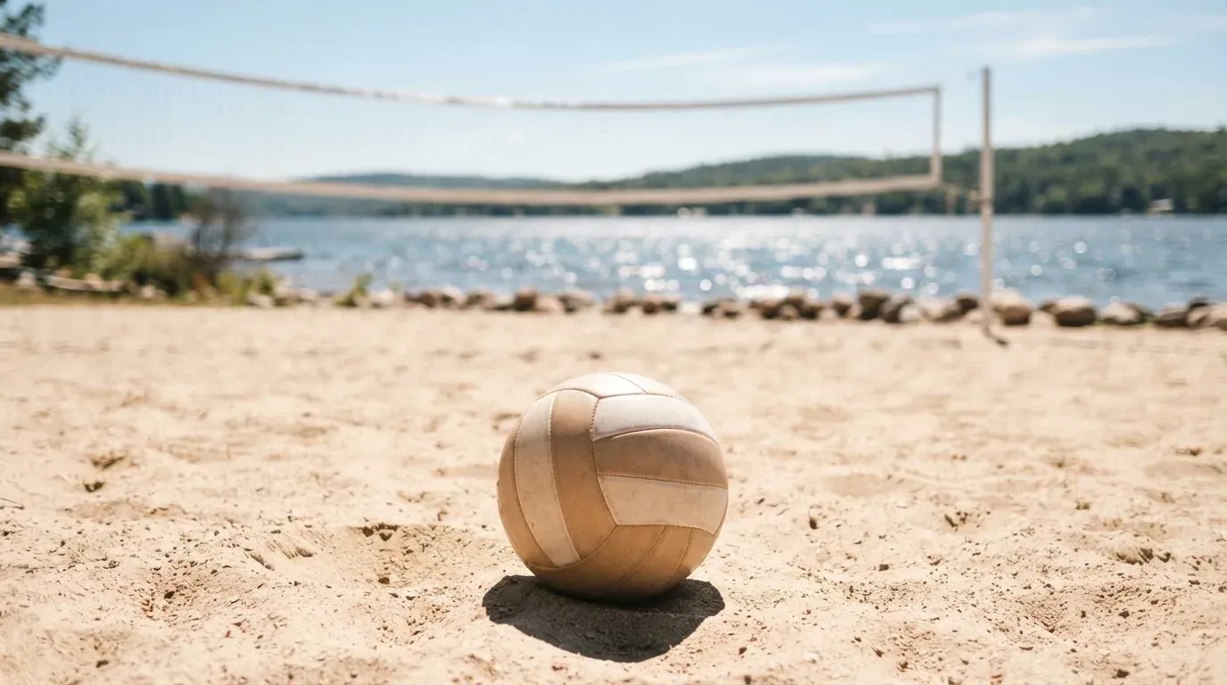 Ein Volleyball liegt auf dem Sand eines Strandfeldes am Seeufer, im Hintergrund glitzert das Wasser.