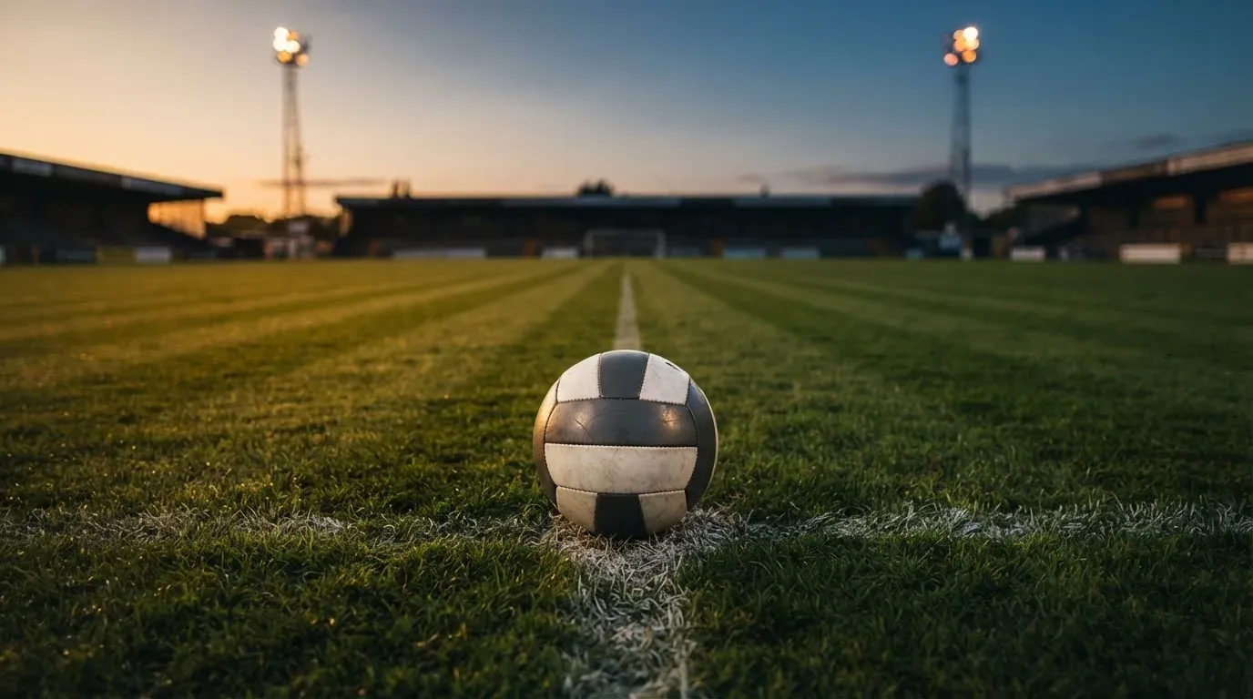 Ein Fussball liegt auf dem Mittelpunkt eines leeren Rasenplatzes, im Hintergrund ragen Flutlichtmasten in die Abenddämmerung