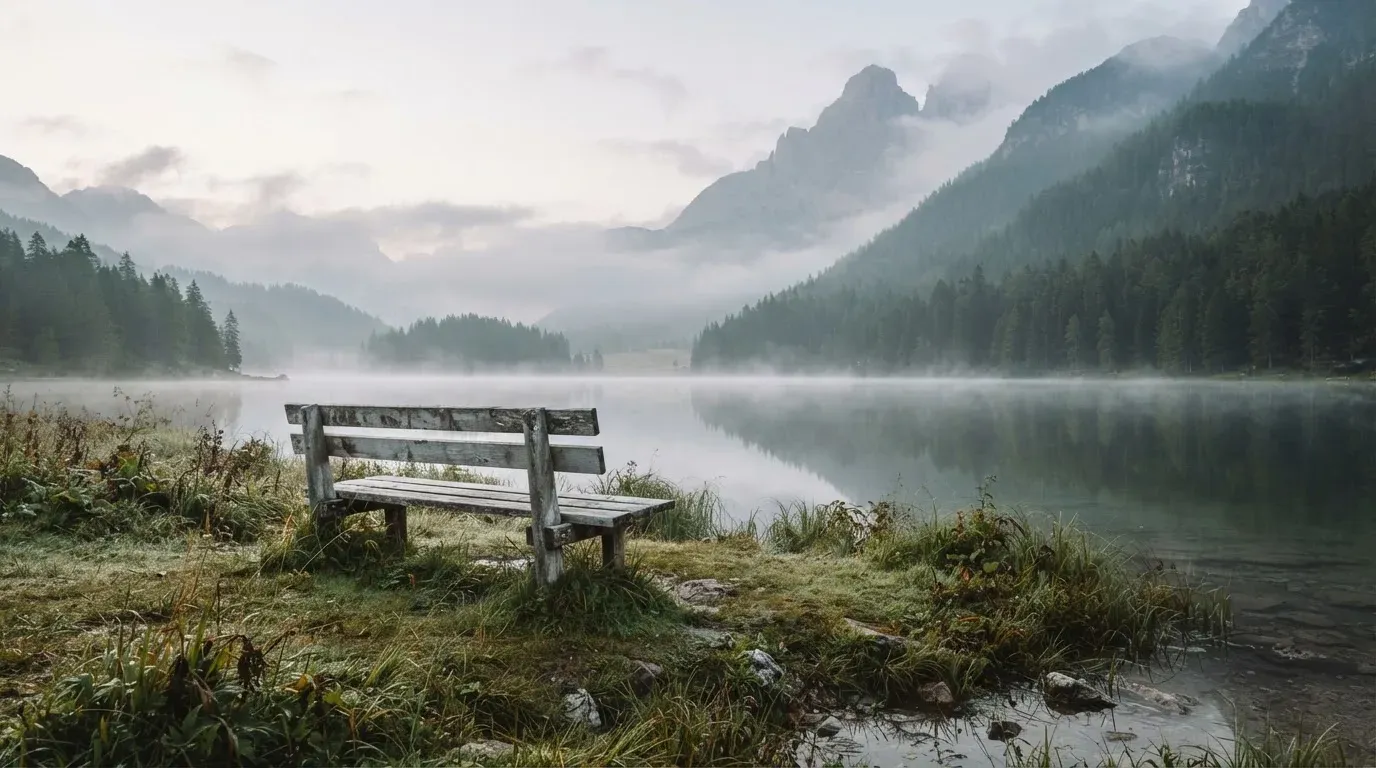 Eine Holzbank am Ufer eines ruhigen Bergsees, am gegenüberliegenden Ufer steigen dunkle Hänge in den Frühnebel