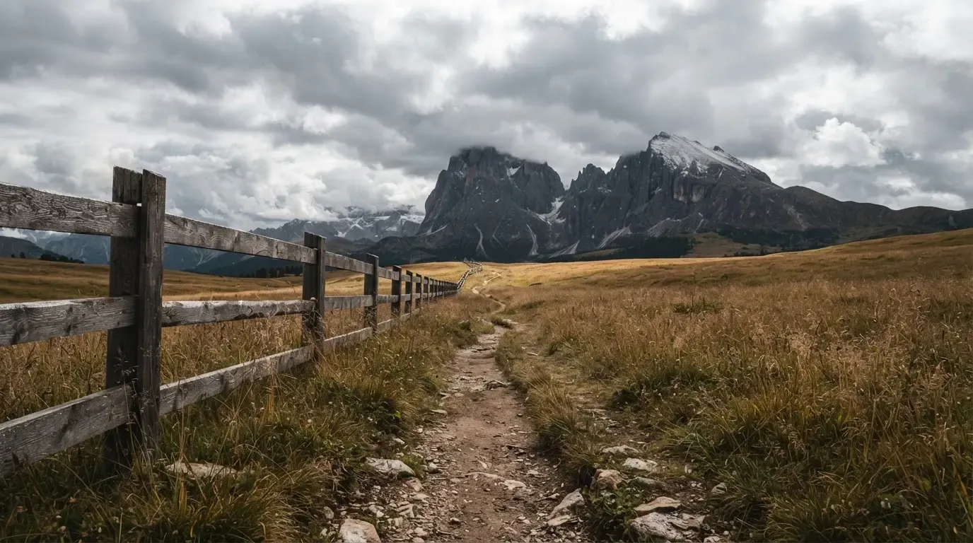 Ein Feldweg führt durch eine Alpwiese mit Holzzaun, im Hintergrund hängen Wolken über einem felsigen Gipfel