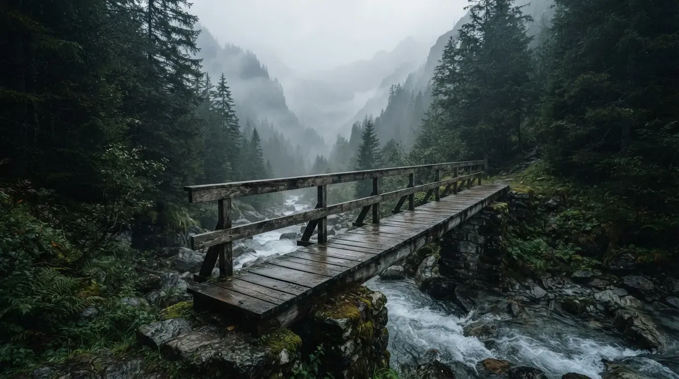 Eine alte Holzbrücke über einen Gebirgsbach, Moos auf den Steinen, Nebelschwaden hängen im Tal