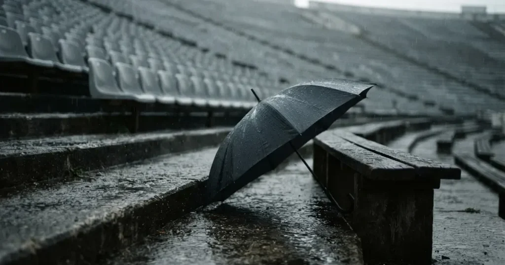 Ein offener Regenschirm lehnt an einer leeren Tribünenbank, Regen fällt auf die Stufen eines Stadions