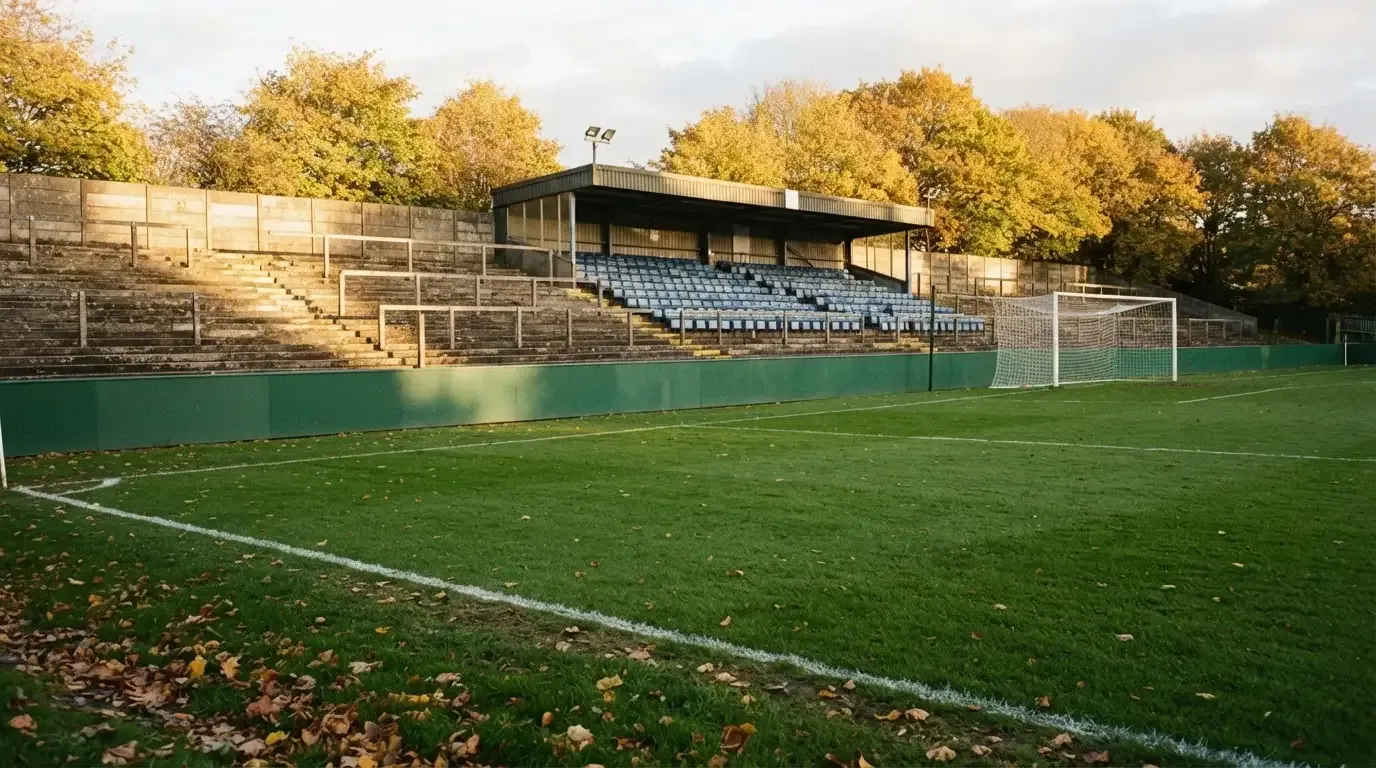 Leere Zuschauerränge eines kleinen Stadions im Herbstlicht, davor ein Rasenplatz mit frisch gezogenen Linien.