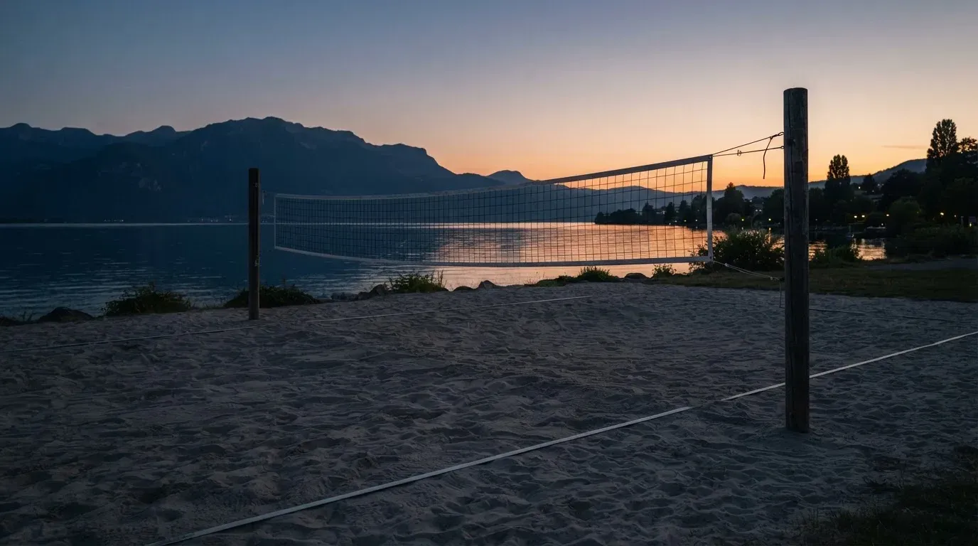 Ein Volleyballnetz steht an einem Seeufer in der Abenddämmerung, der Sand ist leer und das Wasser glatt.