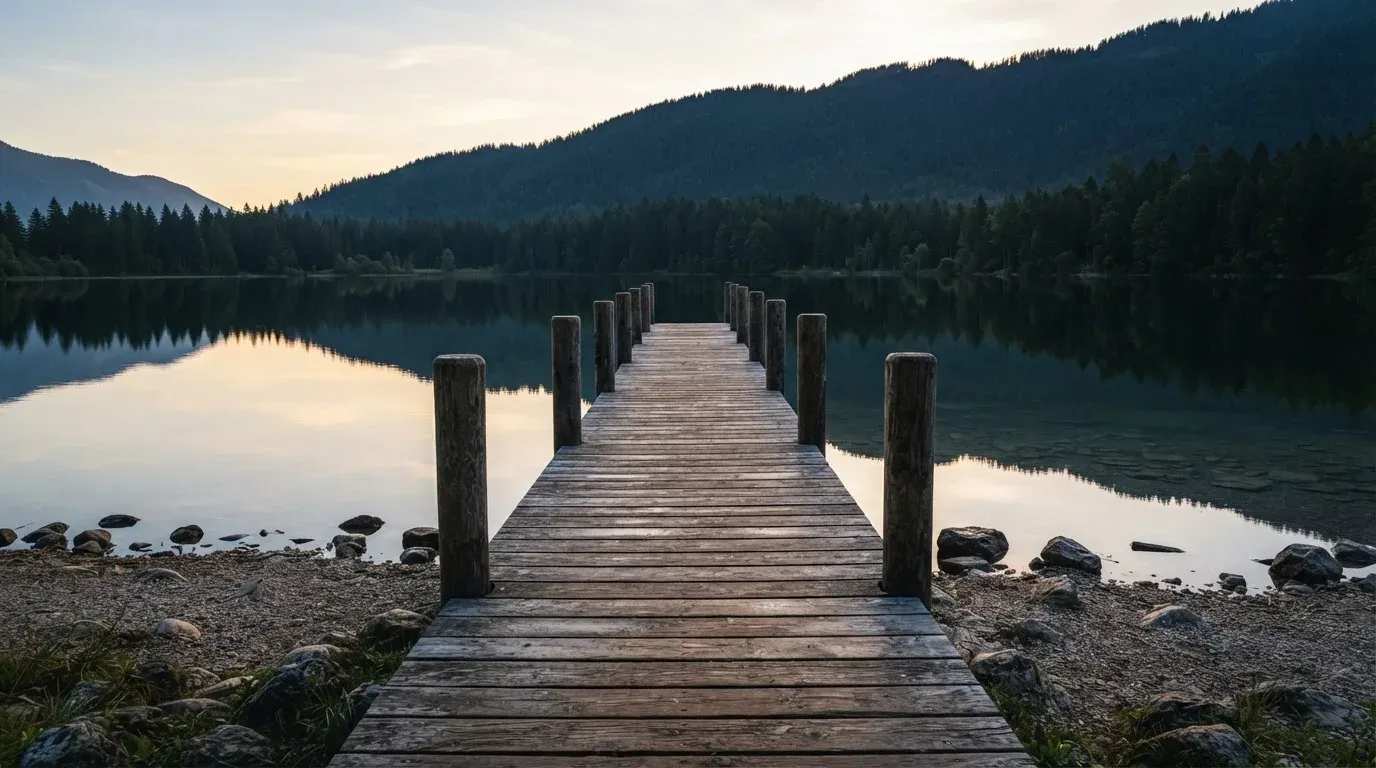 Ein leerer Holzsteg ragt am Abend in einen ruhigen Bergsee, das Wasser spiegelt den Himmel.