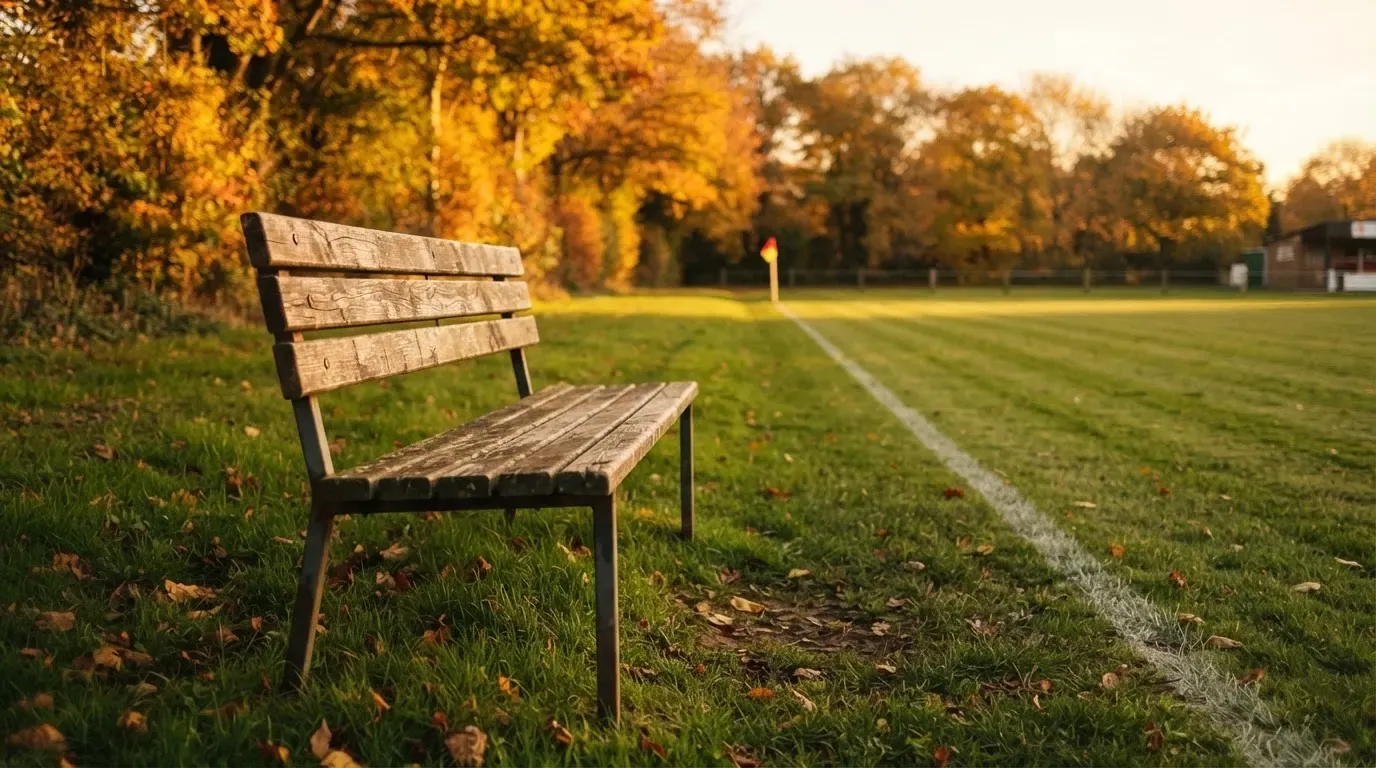 Eine hölzerne Bank steht an der Seitenlinie eines leeren Sportplatzes, dahinter Bäume im Herbstlicht.