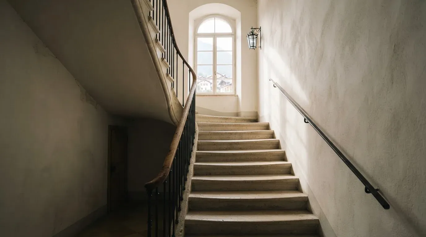 Ein Treppenhaus aus hellem Stein führt in einem Altbau nach oben, Licht fällt durch ein hohes Fenster.